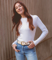 A person with long red hair smiles, wearing the "Wander" white ribbed sweater by Bed Stu, paired with blue jeans and a beige belt that adds whimsical charm, standing in front of a textured wall.