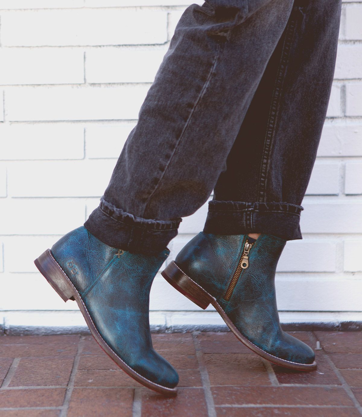 A person wearing Yurisa dark blue leather ankle boots with a side zipper by Bed Stu and black jeans stands on a brick pavement against a white brick wall, showcasing the timeless design and practical elegance of an everyday boot.