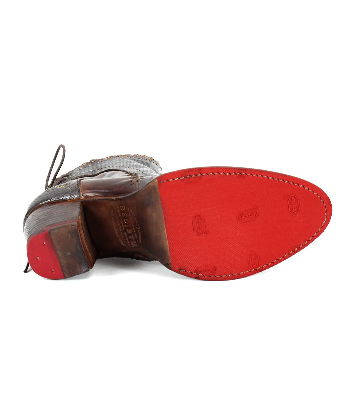 The image shows the bottom view of the Bia, a comfortable leather bootie from Bed Stu, featuring a red sole and visible stitching. The shoe boasts a dark brown upper part and a leather heel with the brand's logo prominently displayed on the sole, emphasizing its handcrafted quality.