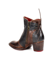 The Bia bootie by Bed Stu is a handcrafted leather piece in brown and black with a side zipper, lace-up detailing at the back, and a chunky heel. It showcases a two-tone design with comfortable red accents inside and on the sole.
