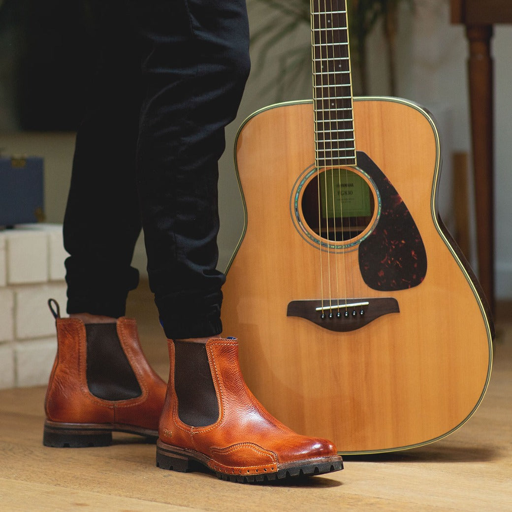 A person standing next to an Brady Trek acoustic guitar, wearing shiny brown Bed Stu chelsea boots with a Vibram outsole on a wooden floor.