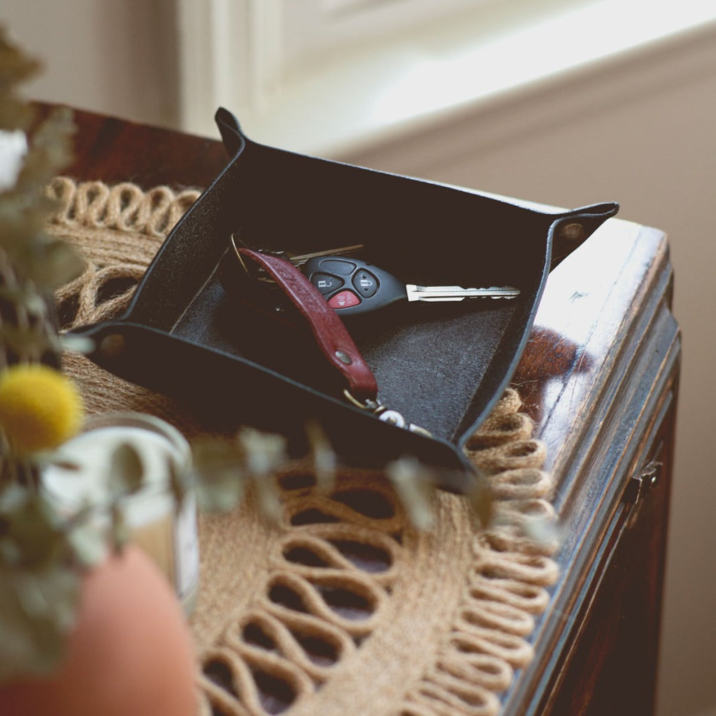 A black Expanse Catchall Tray by Bed Stu holding a set of car keys, a multi-tool knife, and some loose change sits on a wooden surface next to a window.