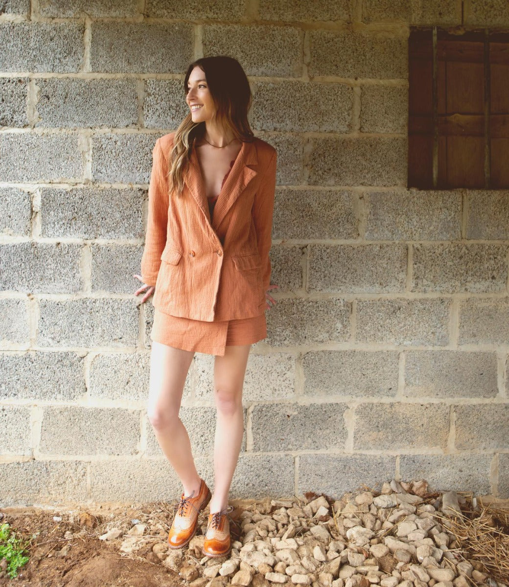 A woman wearing an orange blazer with tan and brown Lita shoes while leaning against a brick wall.