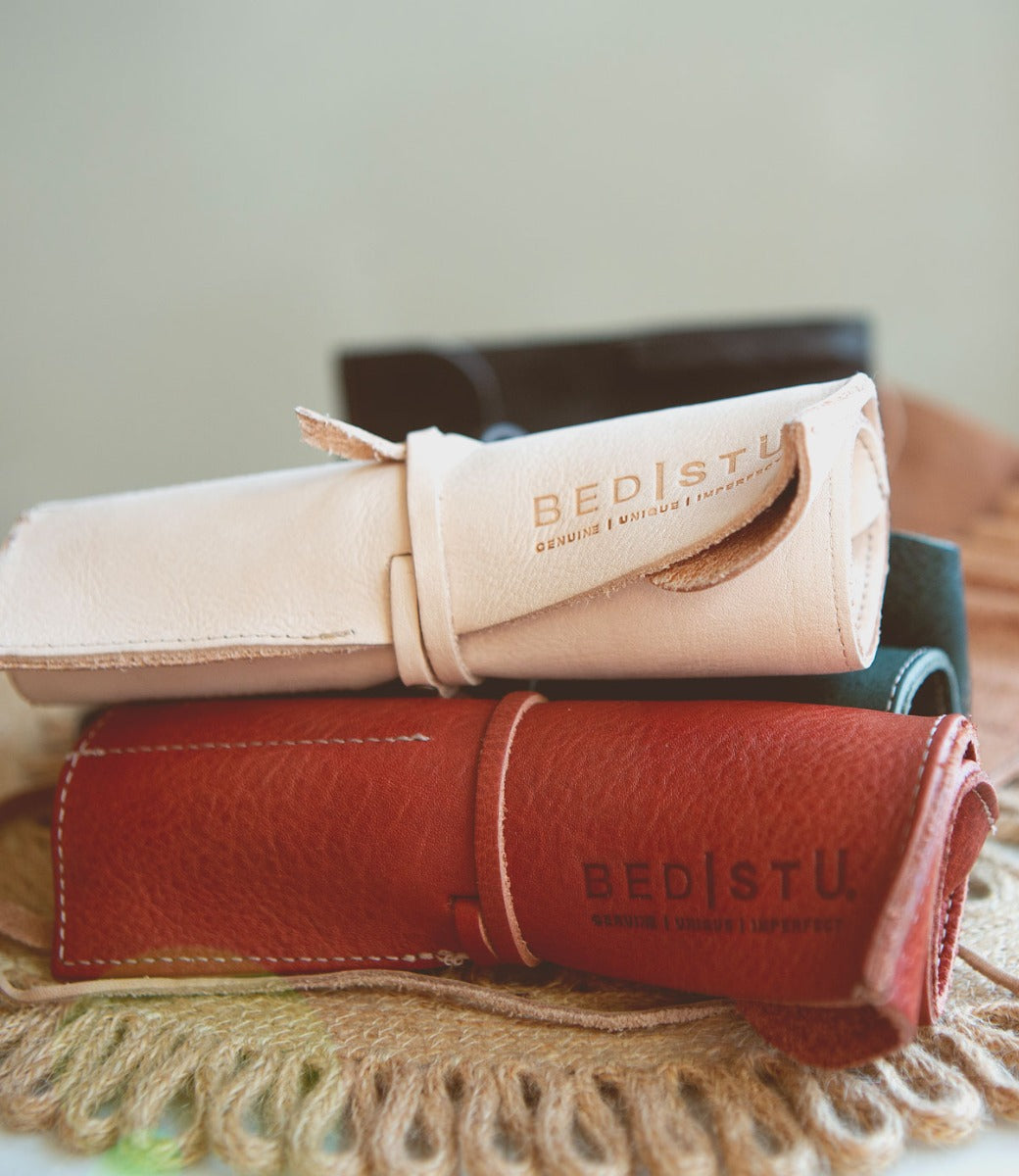Three Prepped Leather Wraps with embossed "Bed Stu" branding are stacked, featuring colors in light pink, black, and red. The artist tools rest on a textured woven surface, evoking the craftsmanship of a Leather Shoppe.